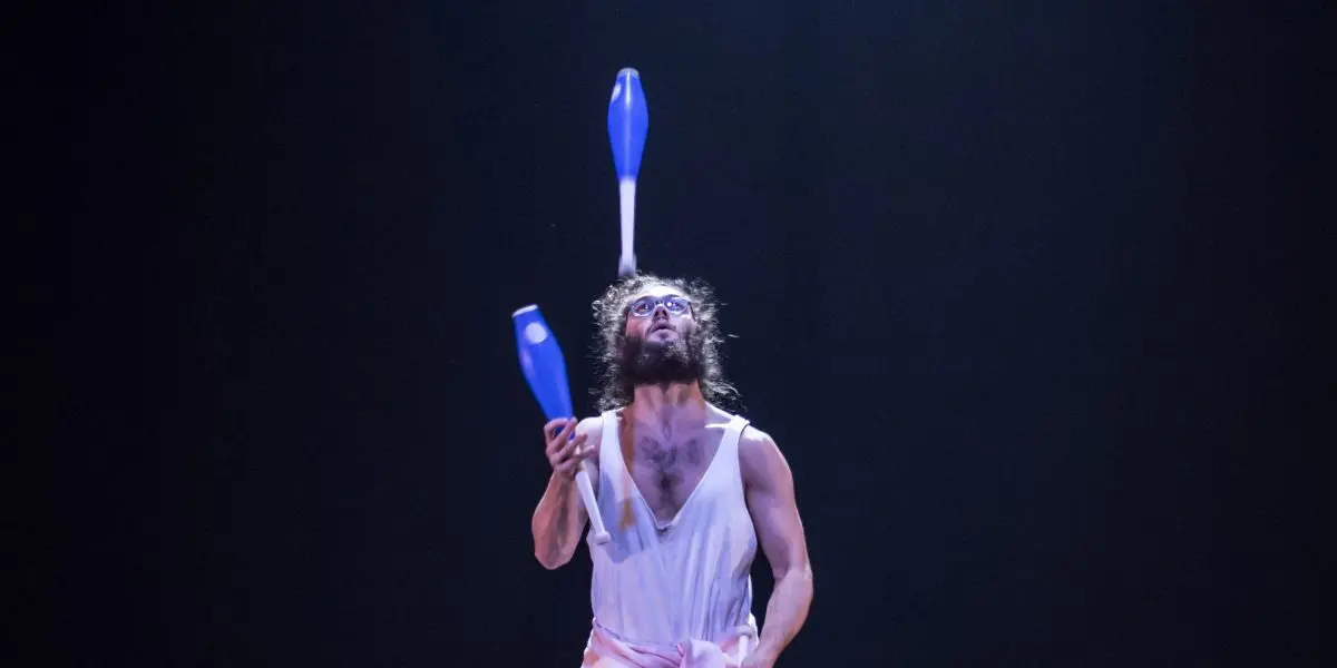 Un jongleur en tenue de sport se tient sur une scène sombre, jonglant habilement avec six massues bleues et blanches sous un projecteur - un spectacle impressionnant qui rappelle les performances dynamiques de Kellin-Quinn Hentoff-Killian.