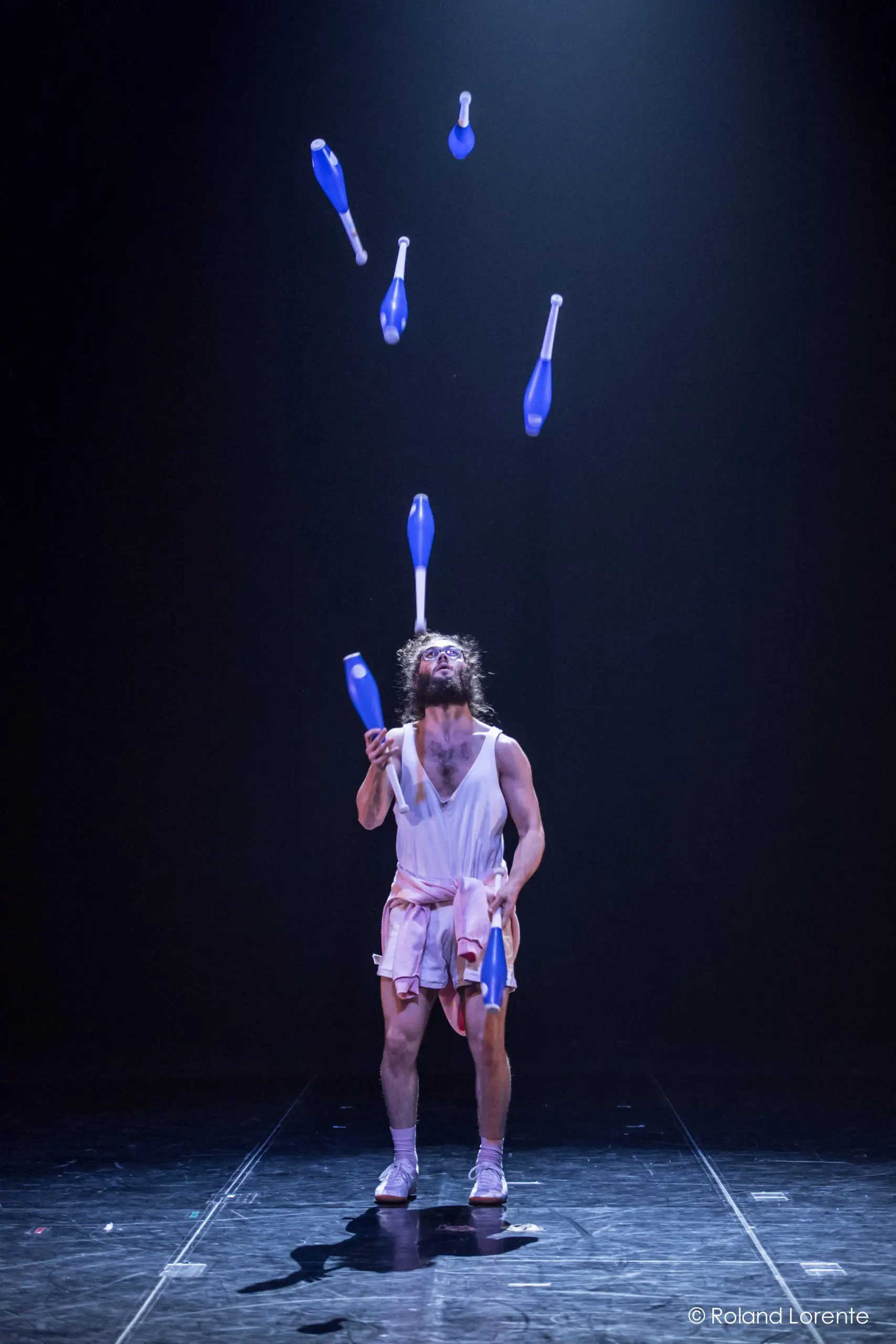 Un jongleur en tenue de sport se tient sur une scène sombre, jonglant habilement avec six massues bleues et blanches sous un projecteur - un spectacle impressionnant qui rappelle les performances dynamiques de Kellin-Quinn Hentoff-Killian.