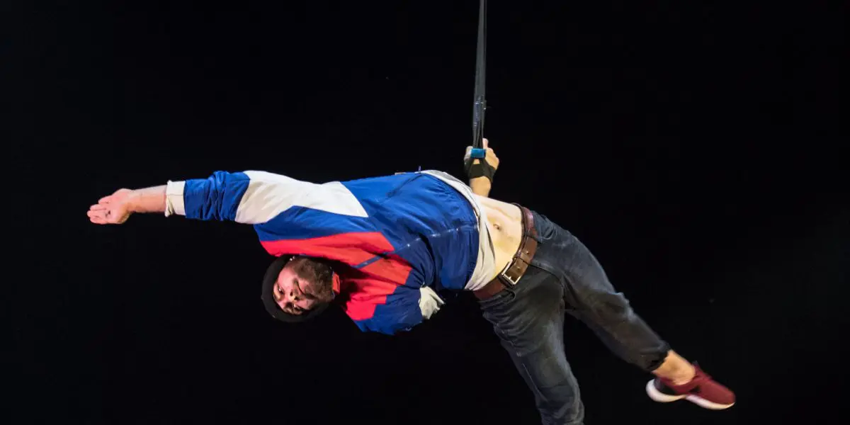Tristan Robquin est suspendu à un harnais, vêtu d'une veste bleue, blanche et rouge sur fond noir, un bras tendu et les jambes pliées, comme s'il flottait ou dansait.