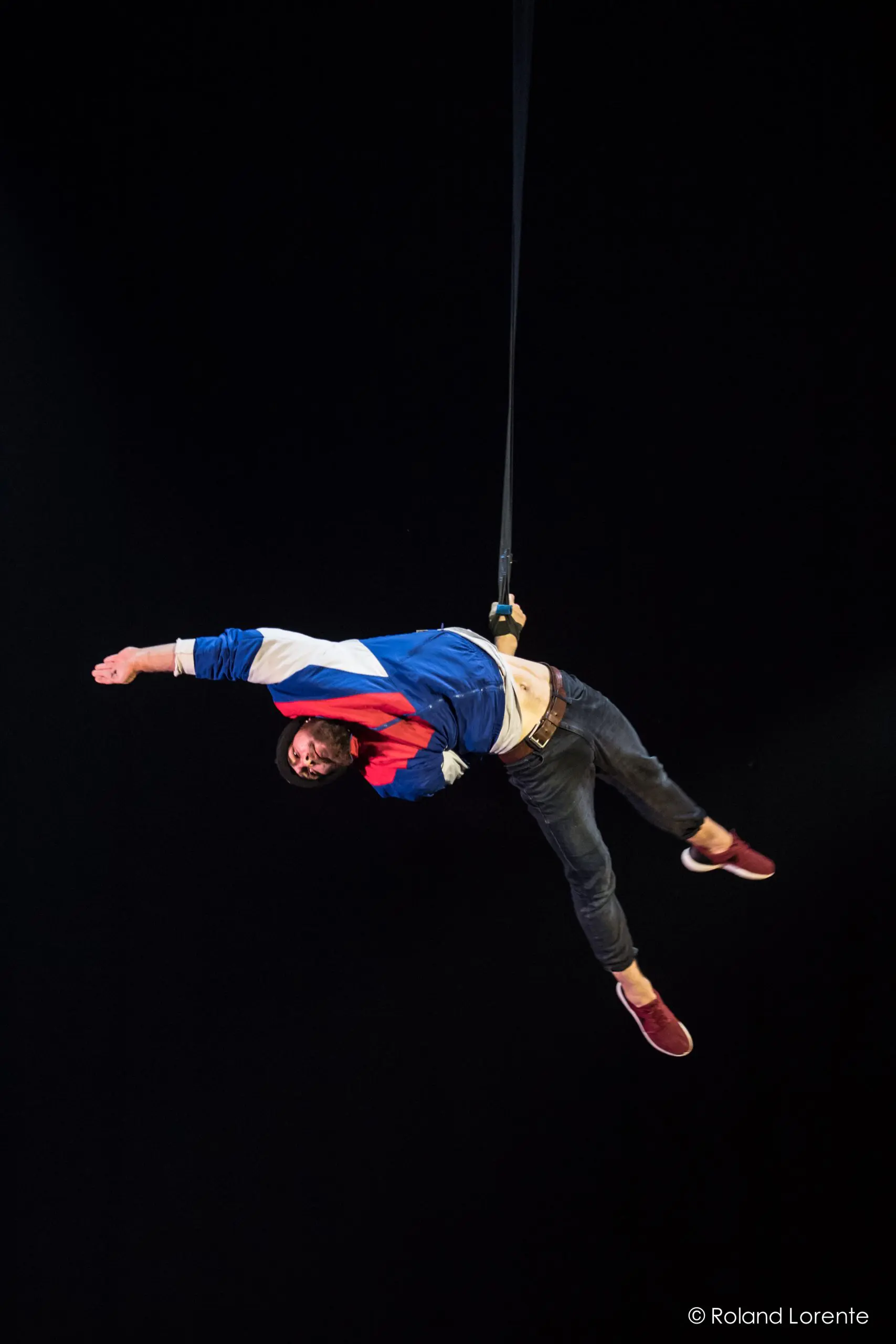 Tristan Robquin est suspendu à un harnais, vêtu d'une veste bleue, blanche et rouge sur fond noir, un bras tendu et les jambes pliées, comme s'il flottait ou dansait.