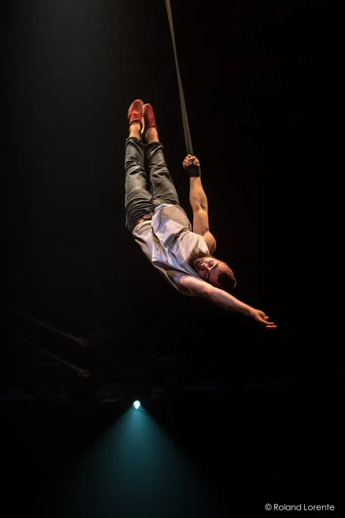 L'artiste aérien Tristan Robquin est suspendu à l'envers par un bras sur une sangle, vêtu d'un débardeur blanc, d'un jean et de chaussures rouges, avec un bras tendu et un projecteur qui brille par en dessous.