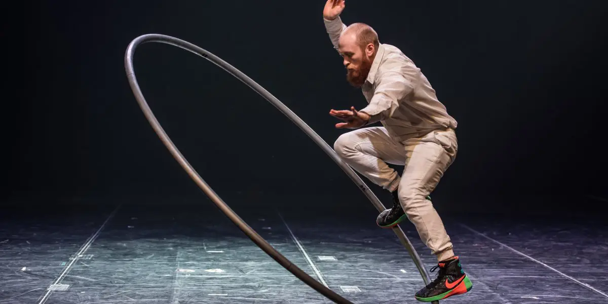 Cooper Smith, vêtu de blanc, se tient habilement en équilibre sur une grande roue Cyr sur une scène sombre. Avec une barbe posée et un bras levé pour garder l'équilibre, sa performance captive le public.