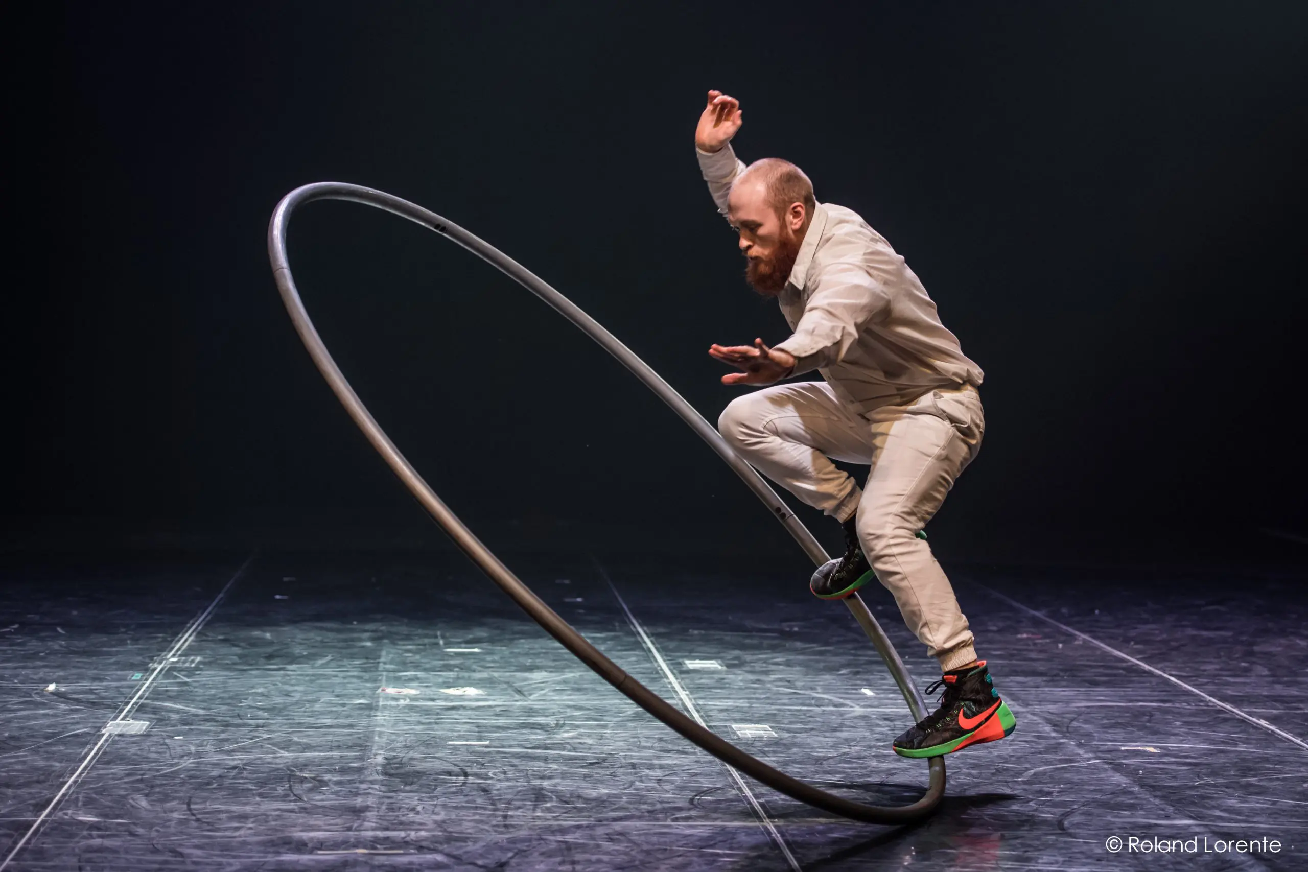 Cooper Smith, vêtu de blanc, se tient habilement en équilibre sur une grande roue Cyr sur une scène sombre. Avec une barbe posée et un bras levé pour garder l'équilibre, sa performance captive le public.