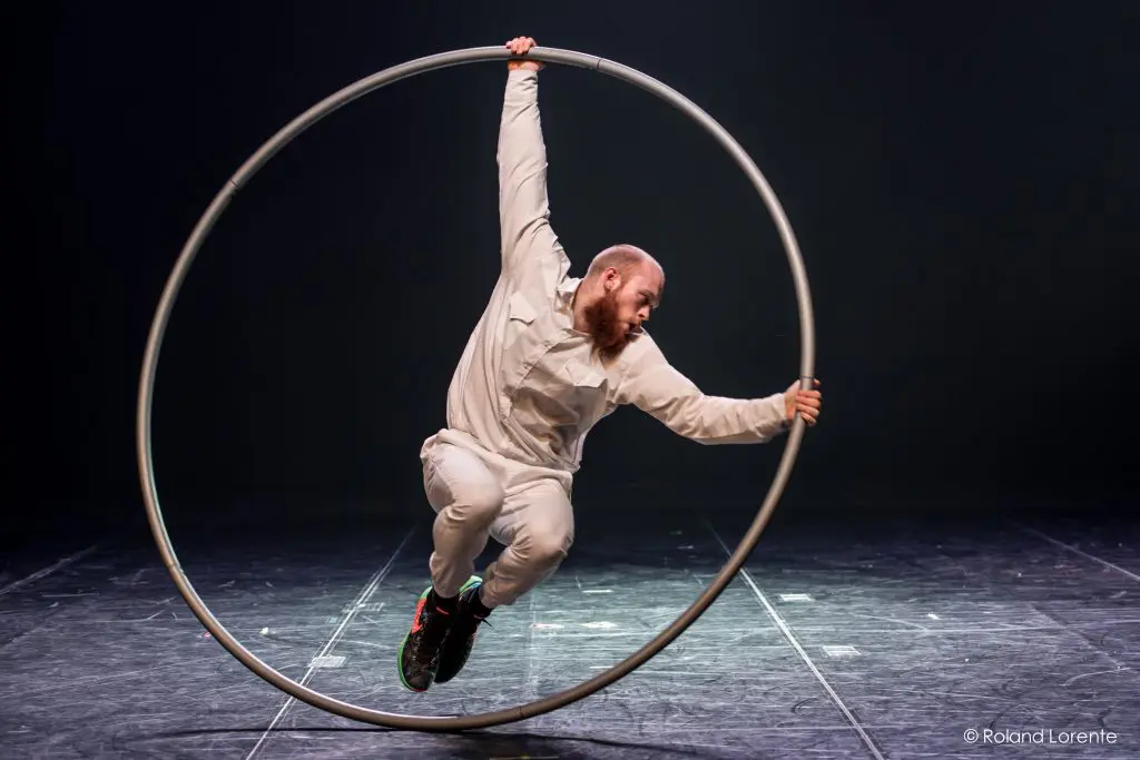 Cooper Smith, vêtu de blanc, se tient en équilibre et tourne à l'intérieur d'une grande roue Cyr en métal sur une scène faiblement éclairée, faisant preuve de force et d'agilité.