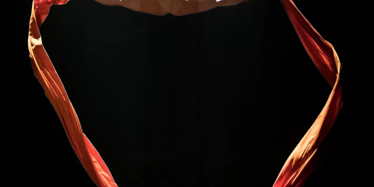 L'artiste aérienne Sabine Van Rensburg, vêtue d'un costume fauve, fait un grand écart au-dessus du sol, suspendue par des rubans de soie rouge et orange et éclairée par un projecteur dans un décor sombre.