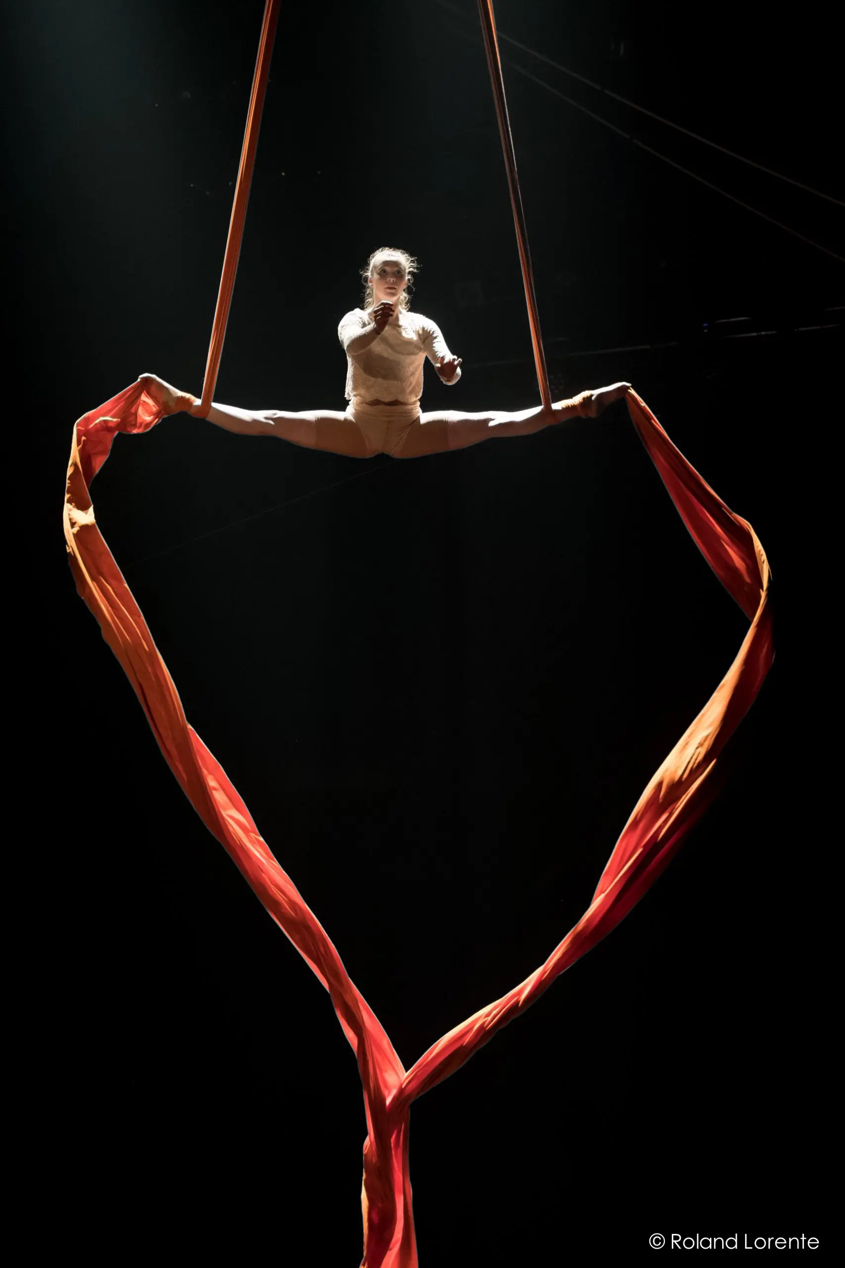 L'artiste aérienne Sabine Van Rensburg, vêtue d'un costume fauve, fait un grand écart au-dessus du sol, suspendue par des rubans de soie rouge et orange et éclairée par un projecteur dans un décor sombre.