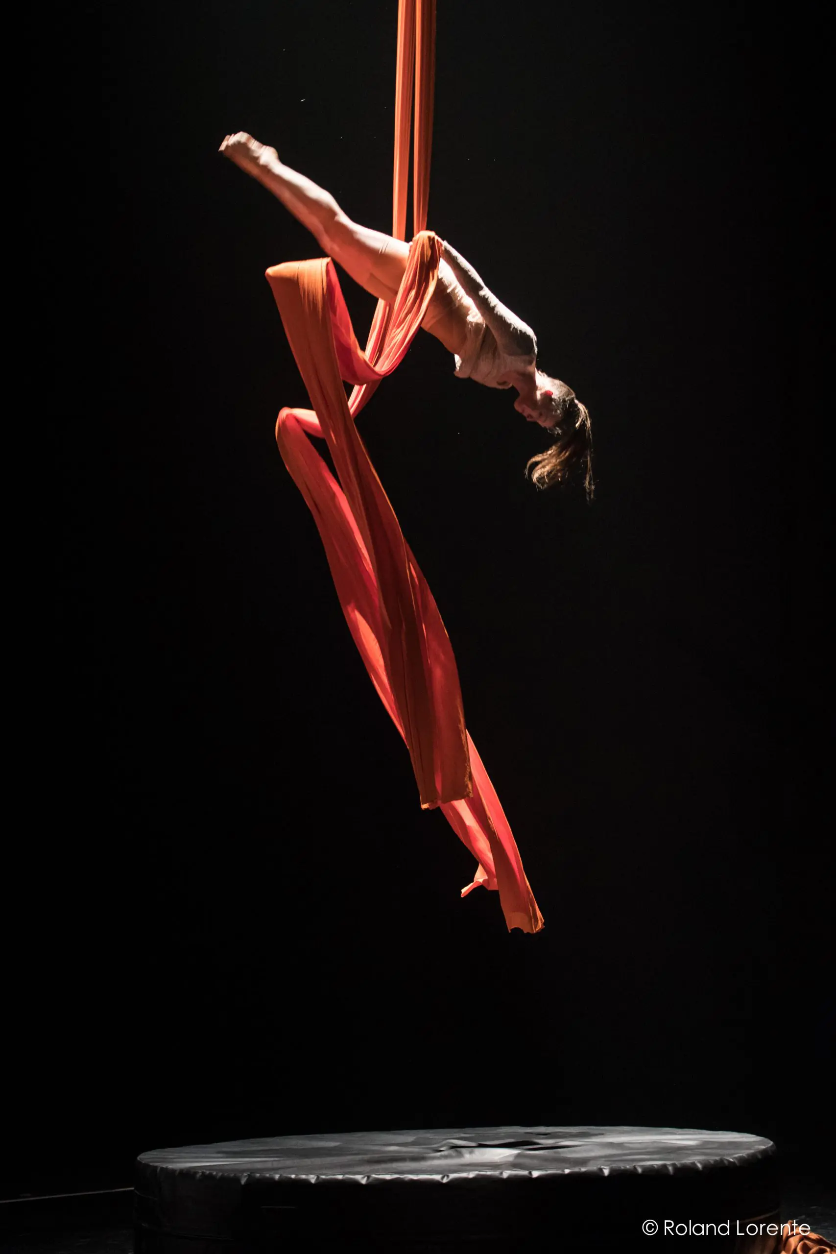 L'artiste aérienne Sabine Van Rensburg est suspendue en l'air sur des soies orange, se balançant gracieusement au-dessus d'un tapis de scène rond et noir, dans un décor sombre.