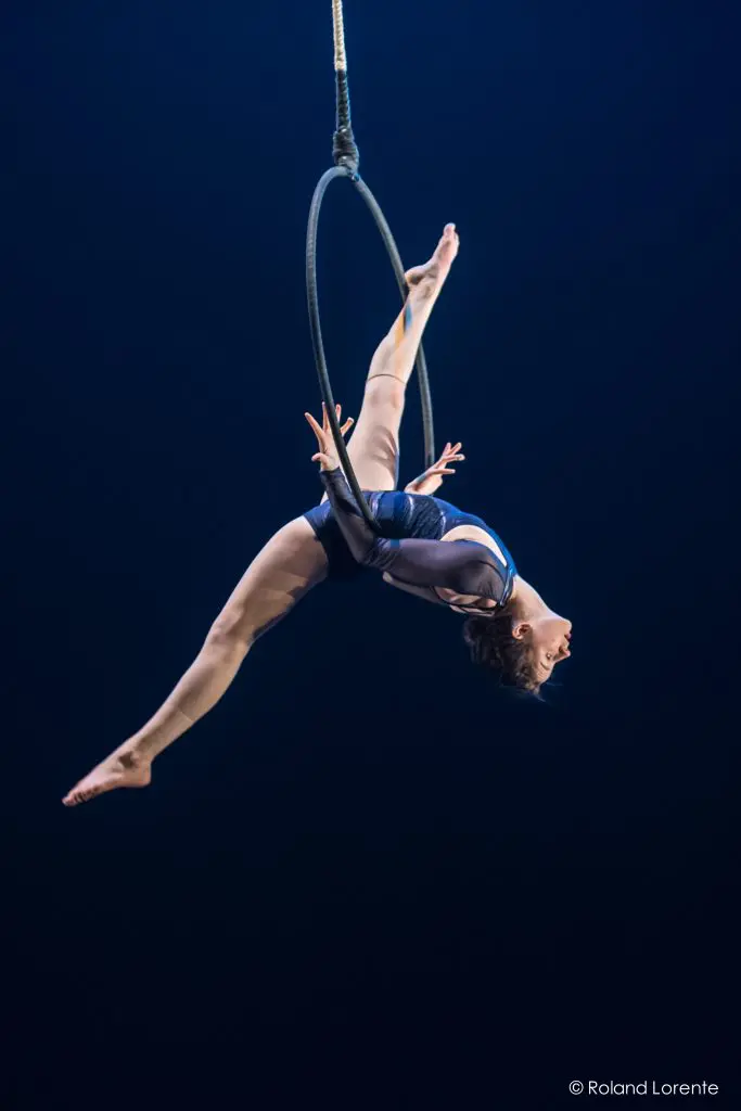 L'artiste de cirque Rae Failing exécute un numéro aérien, se suspend la tête en bas et pose gracieusement sur un cerceau suspendu sur un fond bleu foncé.