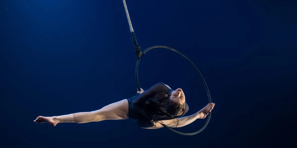 Une voltigeuse, Rae Failing, se produit sur un cerceau suspendu, étendant gracieusement ses bras et ses jambes sur un fond bleu foncé, éclairé par la lumière de la scène.