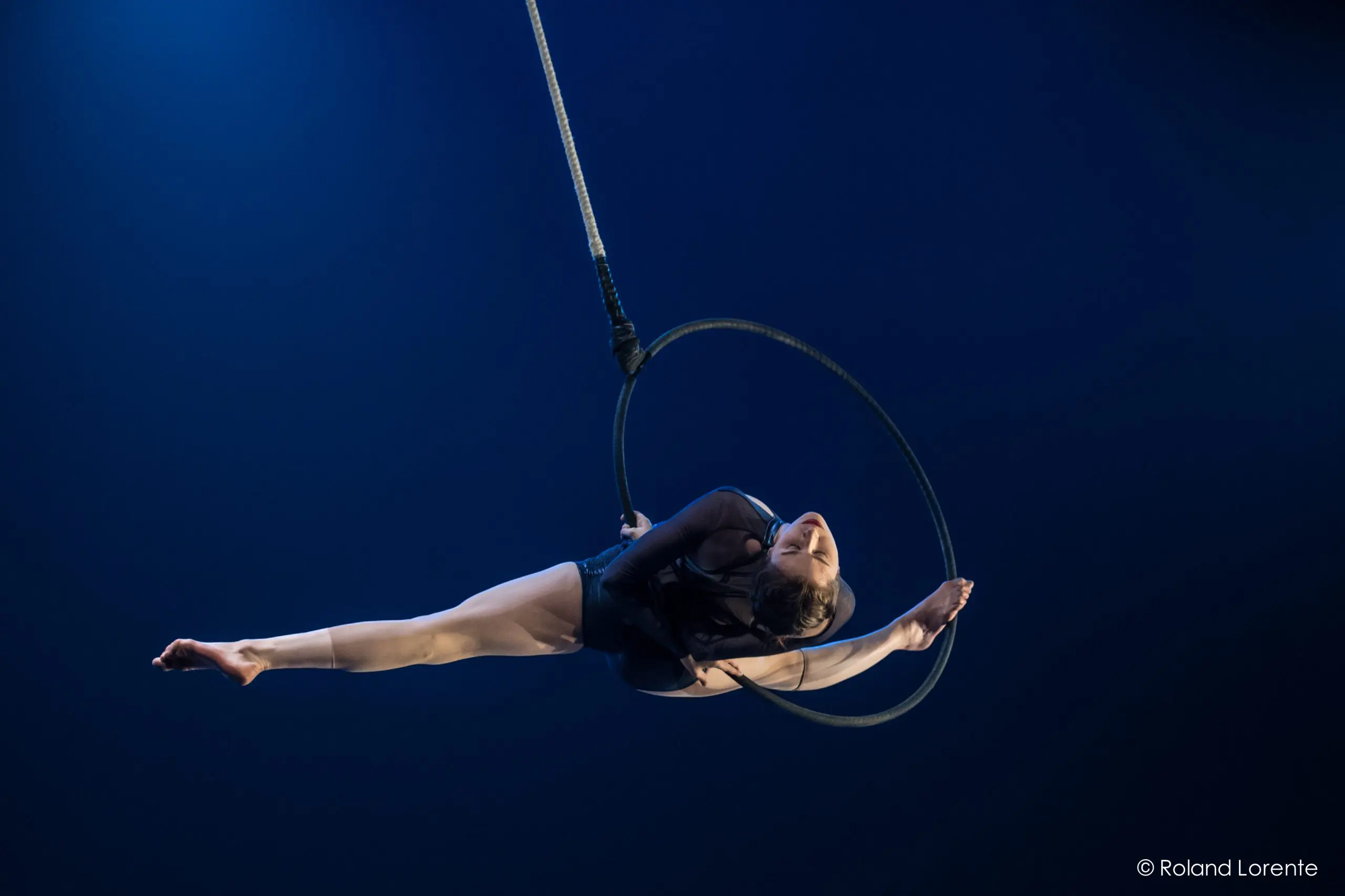 Une voltigeuse, Rae Failing, se produit sur un cerceau suspendu, étendant gracieusement ses bras et ses jambes sur un fond bleu foncé, éclairé par la lumière de la scène.