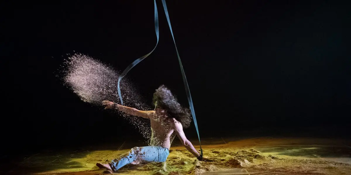 L'interprète Ricardo Casteneyra, aux cheveux longs, est assis sur le sol, dispersant du sable de manière spectaculaire avec un bras tandis qu'un ruban noir tourbillonne au-dessus, éclairé par les lumières de la scène sur un fond sombre.