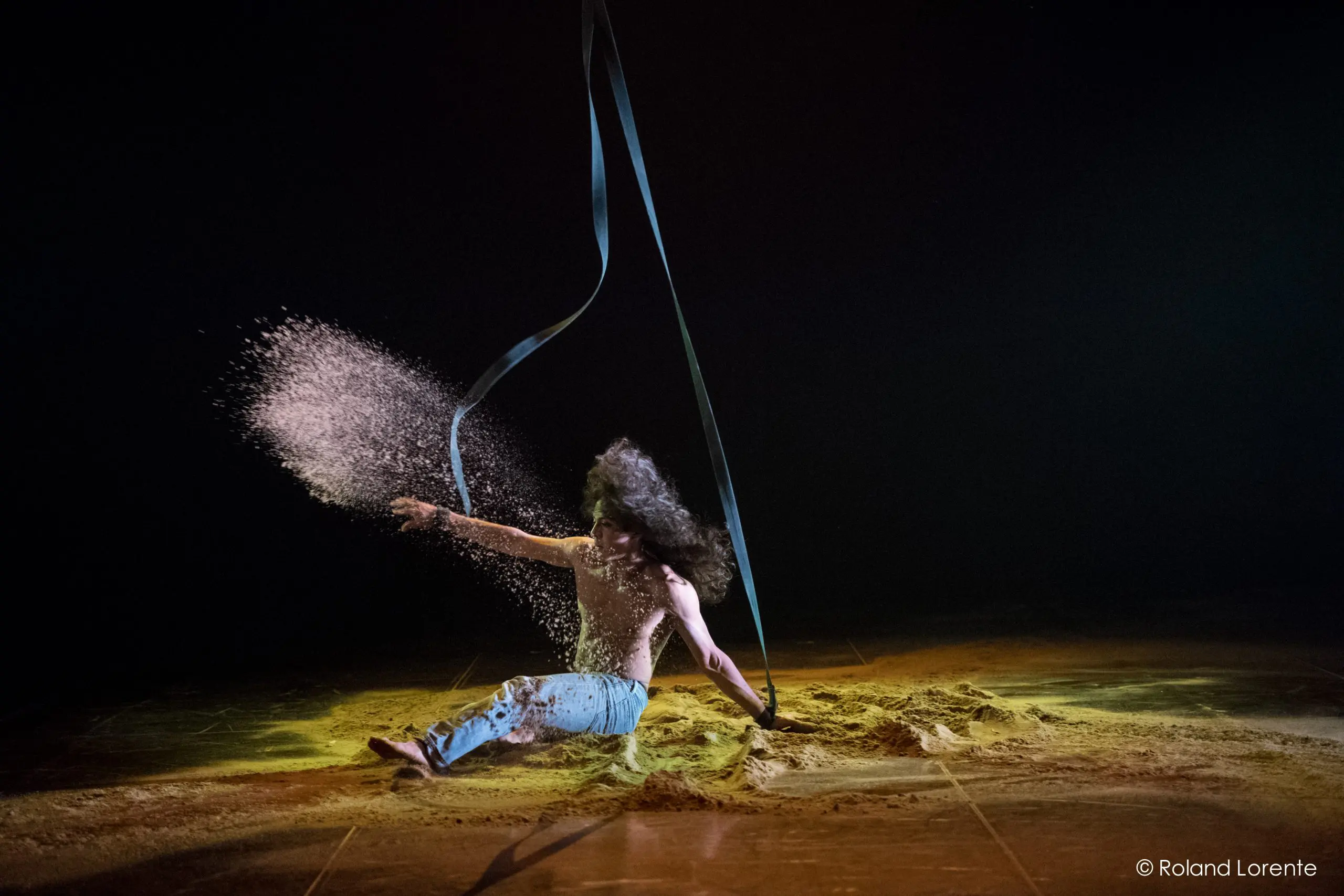 L'interprète Ricardo Casteneyra, aux cheveux longs, est assis sur le sol, dispersant du sable de manière spectaculaire avec un bras tandis qu'un ruban noir tourbillonne au-dessus, éclairé par les lumières de la scène sur un fond sombre.