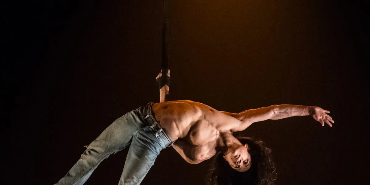 L'acrobate aérien Ricardo Casteneyra, cheveux longs et torse nu, vêtu d'un jean, se produit suspendu à une sangle sous un projecteur sur un fond sombre.