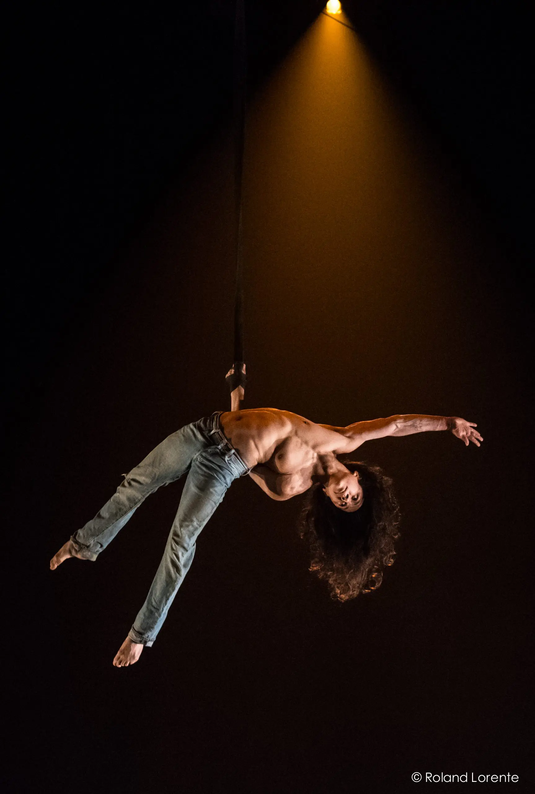L'acrobate aérien Ricardo Casteneyra, cheveux longs et torse nu, vêtu d'un jean, se produit suspendu à une sangle sous un projecteur sur un fond sombre.