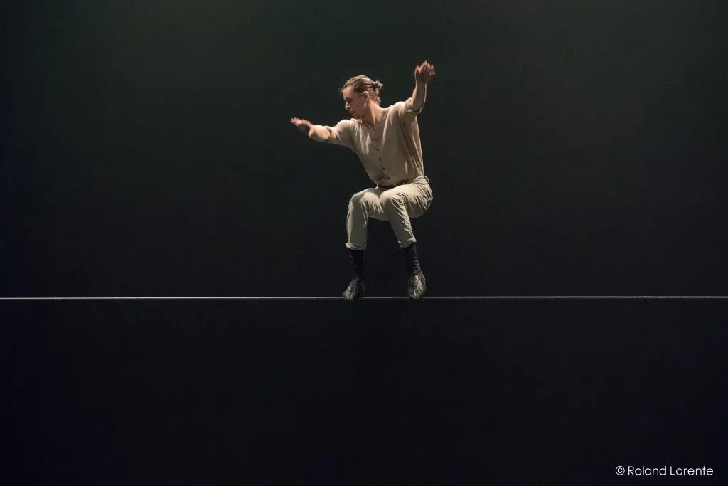 Une personne portant des vêtements clairs et des bottes noires se tient en équilibre sur une corde raide sur un fond sombre, les bras tendus pour assurer sa stabilité.