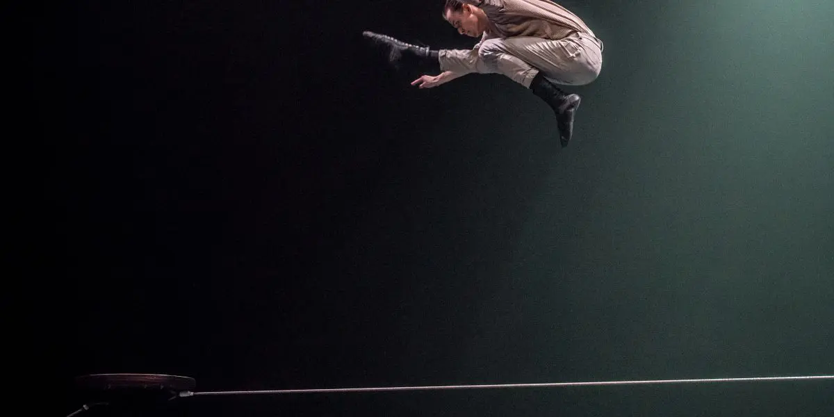 Un artiste saute au-dessus d'une corde raide sous les projecteurs, les jambes étendues et les bras rentrés, sur un fond sombre. Une plate-forme ronde et le fil tendu sont visibles en dessous.