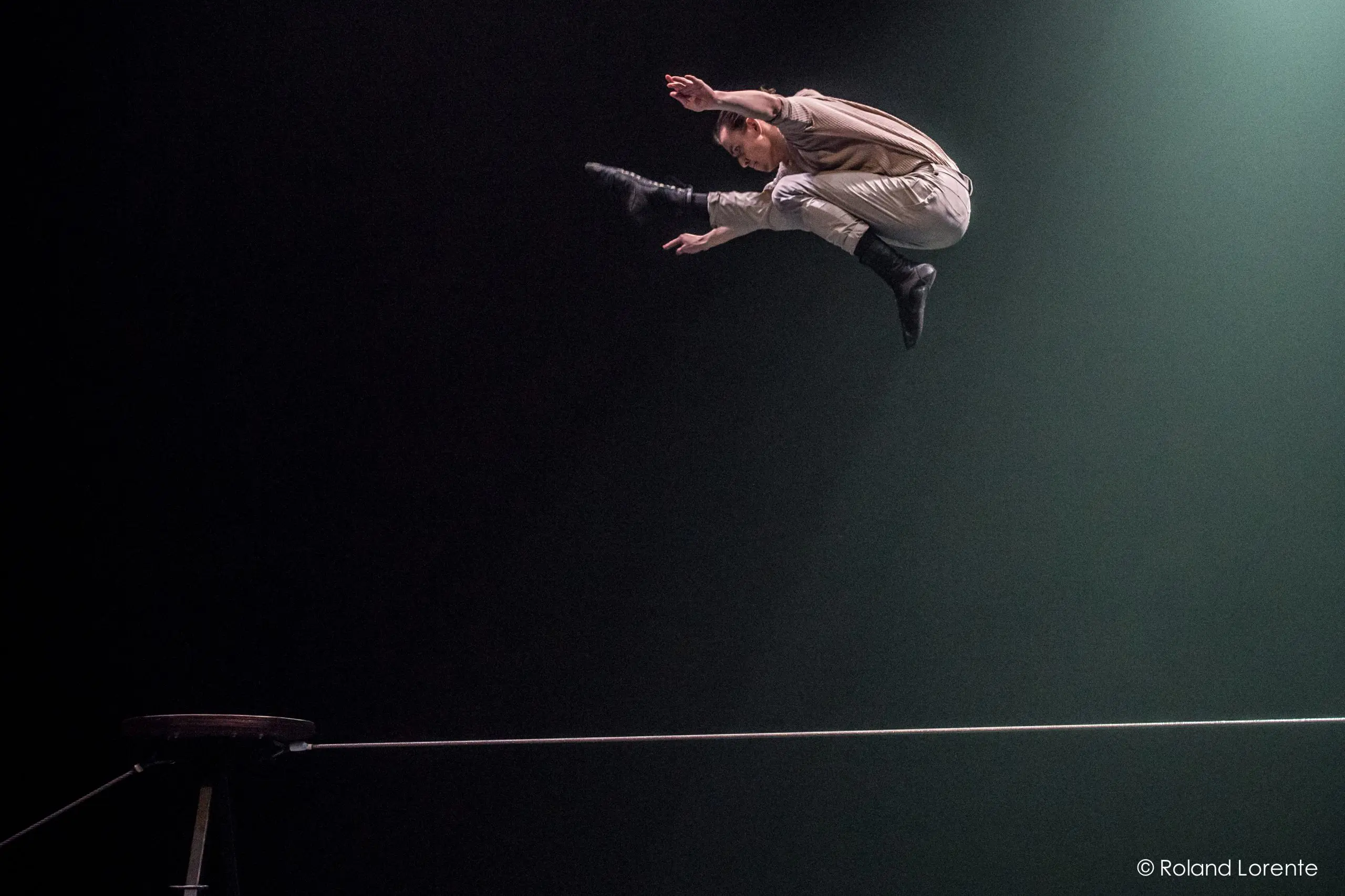 Un artiste saute au-dessus d'une corde raide sous les projecteurs, les jambes étendues et les bras rentrés, sur un fond sombre. Une plate-forme ronde et le fil tendu sont visibles en dessous.