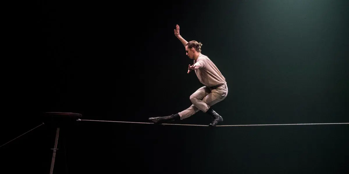 Une personne se tient habilement en équilibre sur une corde raide sous les projecteurs, un genou plié et les bras tendus pour assurer sa stabilité. L'arrière-plan est sombre, ce qui accentue le caractère dramatique de la scène.