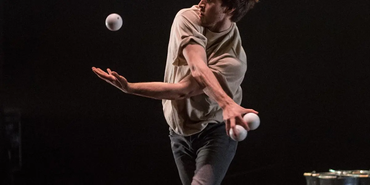 Un homme pieds nus, vêtu d'une chemise beige et d'un pantalon sombre, jongle avec quatre balles blanches sur une scène sombre. Il est capturé en plein mouvement avec une expression concentrée. Un seau en métal est visible à l'arrière-plan. Cette scène dynamique met en scène l'artiste Philippe Dupuis.
