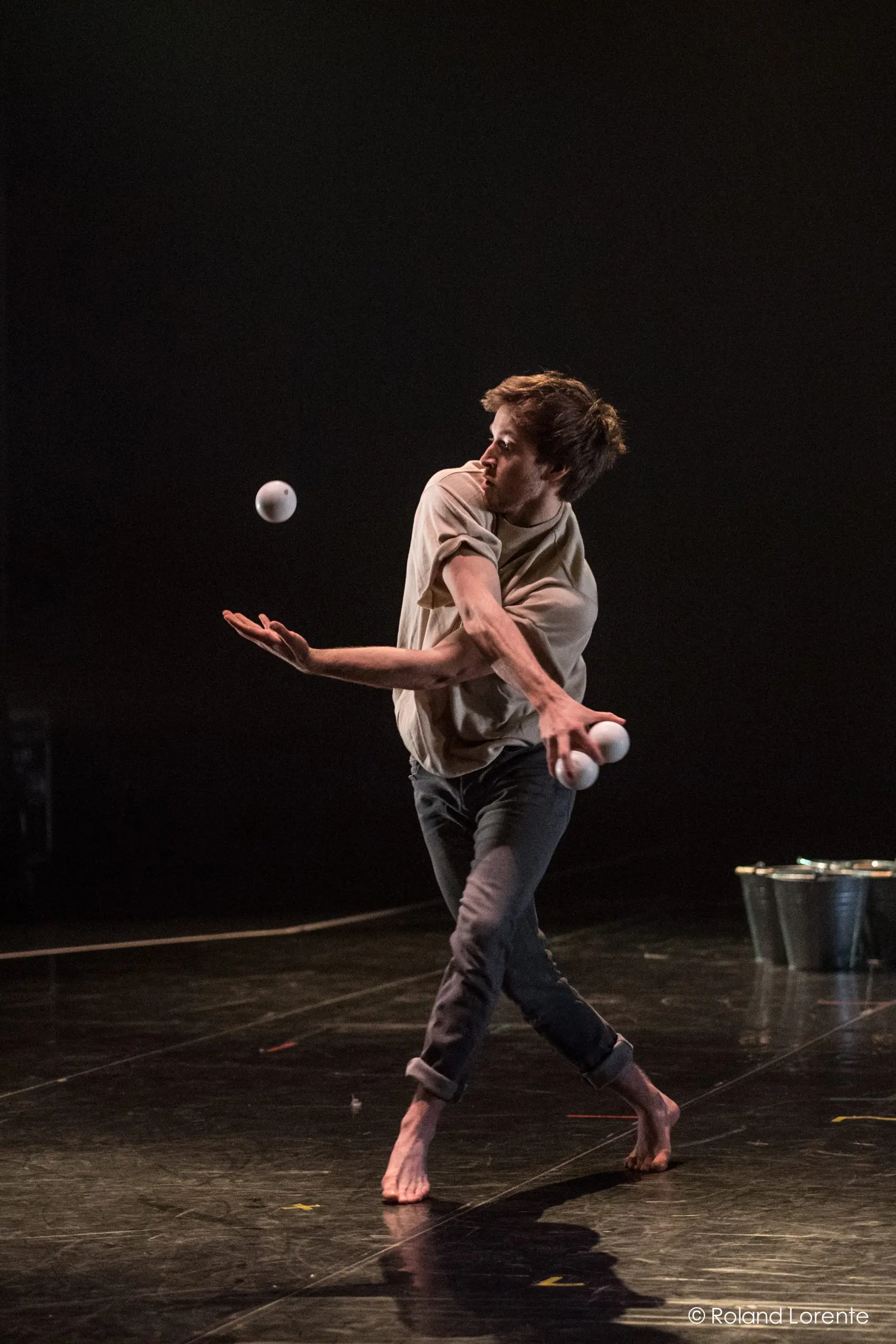 Un homme pieds nus, vêtu d'une chemise beige et d'un pantalon sombre, jongle avec quatre balles blanches sur une scène sombre. Il est capturé en plein mouvement avec une expression concentrée. Un seau en métal est visible à l'arrière-plan. Cette scène dynamique met en scène l'artiste Philippe Dupuis.