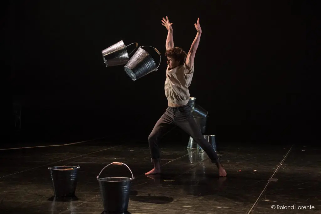 Un danseur pieds nus en tenue décontractée lève les bras sur scène, avec trois seaux métalliques en l'air autour de lui et d'autres seaux sur le sol, sous un éclairage dramatique dans le spectacle captivant de Philippe Dupuis.
