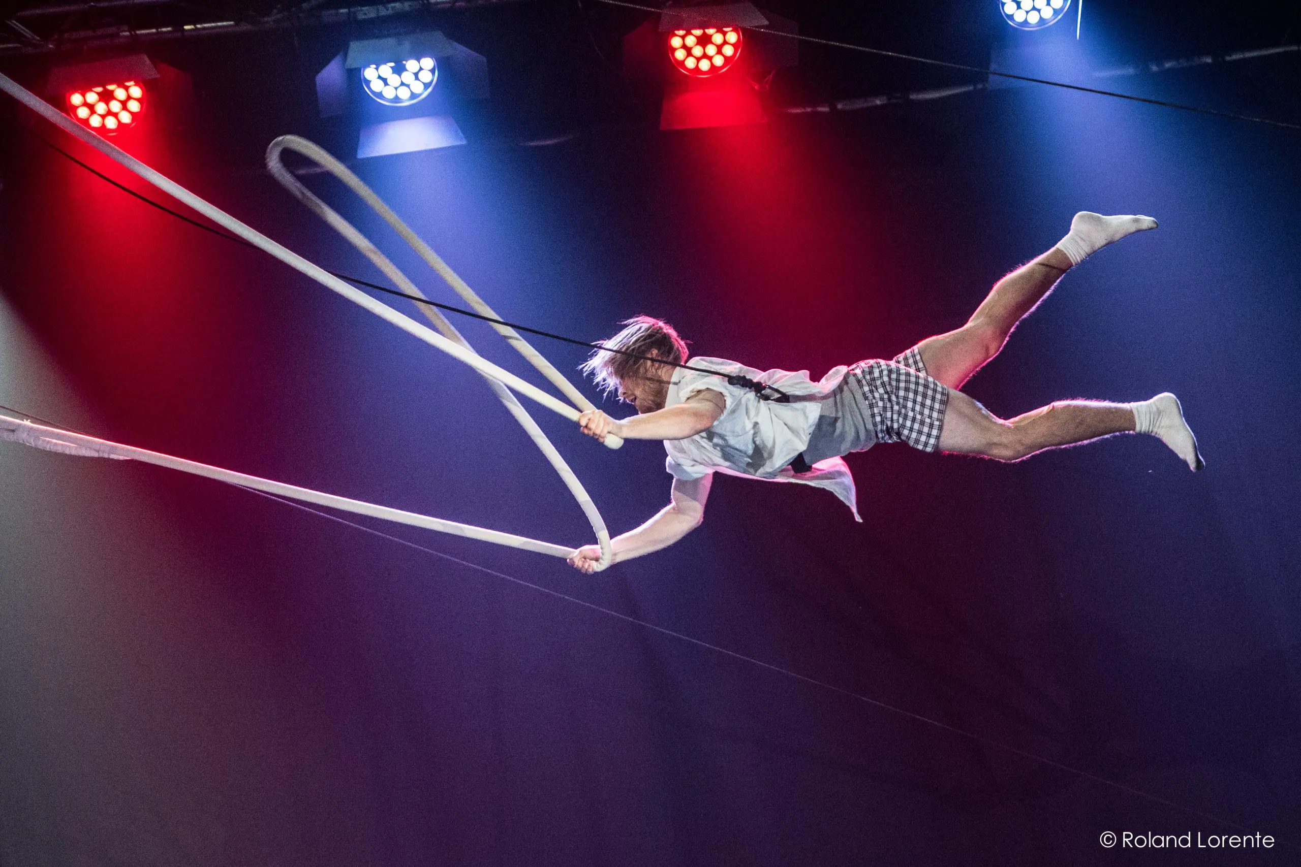 Un artiste de cirque, peut-être Nathan Jones, tient en l'air de longs rubans blancs, vêtu d'une tenue à carreaux et de chaussettes blanches, sous un éclairage de scène rouge et bleu spectaculaire.