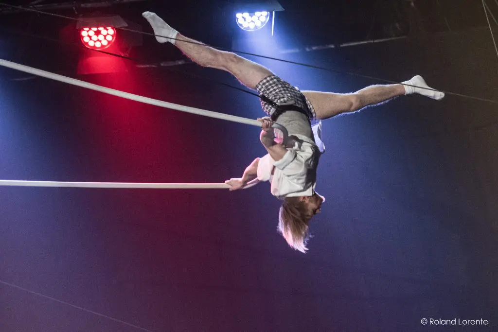 Nathan Jones, un artiste de cirque vêtu d'un short à carreaux et d'une chemise blanche, se suspend la tête en bas et fend les jambes sur une corde raide sous les lumières de la scène, faisant preuve d'une force et d'une souplesse remarquables.