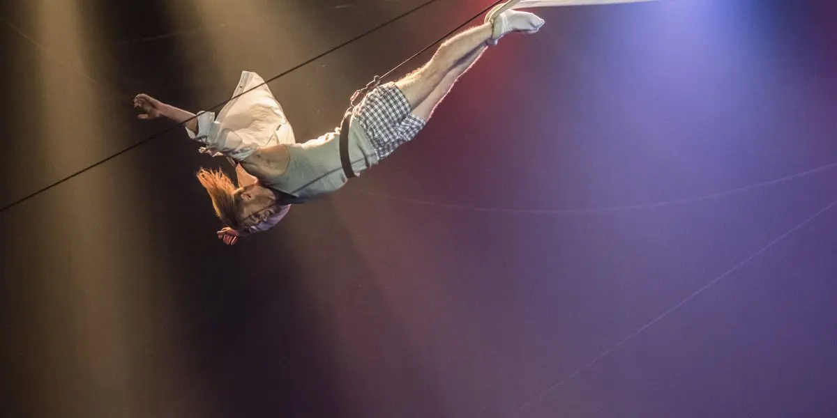 Nathan Jones, vêtu d'une chemise blanche et d'un short à carreaux, est suspendu à l'envers à un fil de fer pendant un numéro d'acrobatie, éclairé par une lumière de scène spectaculaire.