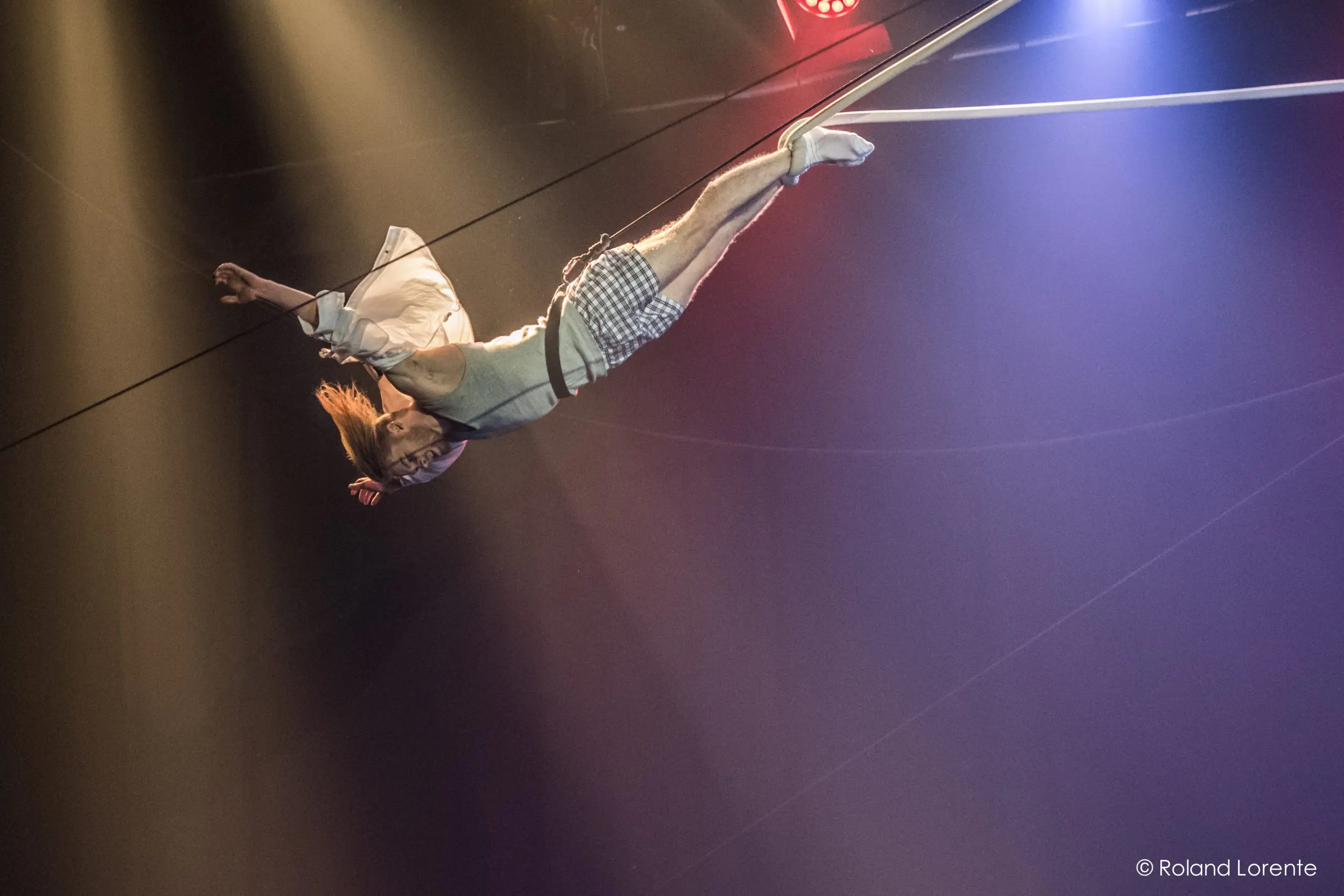Nathan Jones, vêtu d'une chemise blanche et d'un short à carreaux, est suspendu à l'envers à un fil de fer pendant un numéro d'acrobatie, éclairé par une lumière de scène spectaculaire.