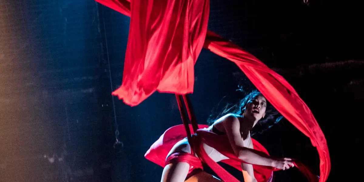 Une artiste aérienne, rappelant Mizuki Shinagawa, est gracieusement suspendue dans les airs, enveloppée dans des rubans de soie rouge fluides, tandis qu'un éclairage de scène dramatique l'illumine sur un fond sombre.