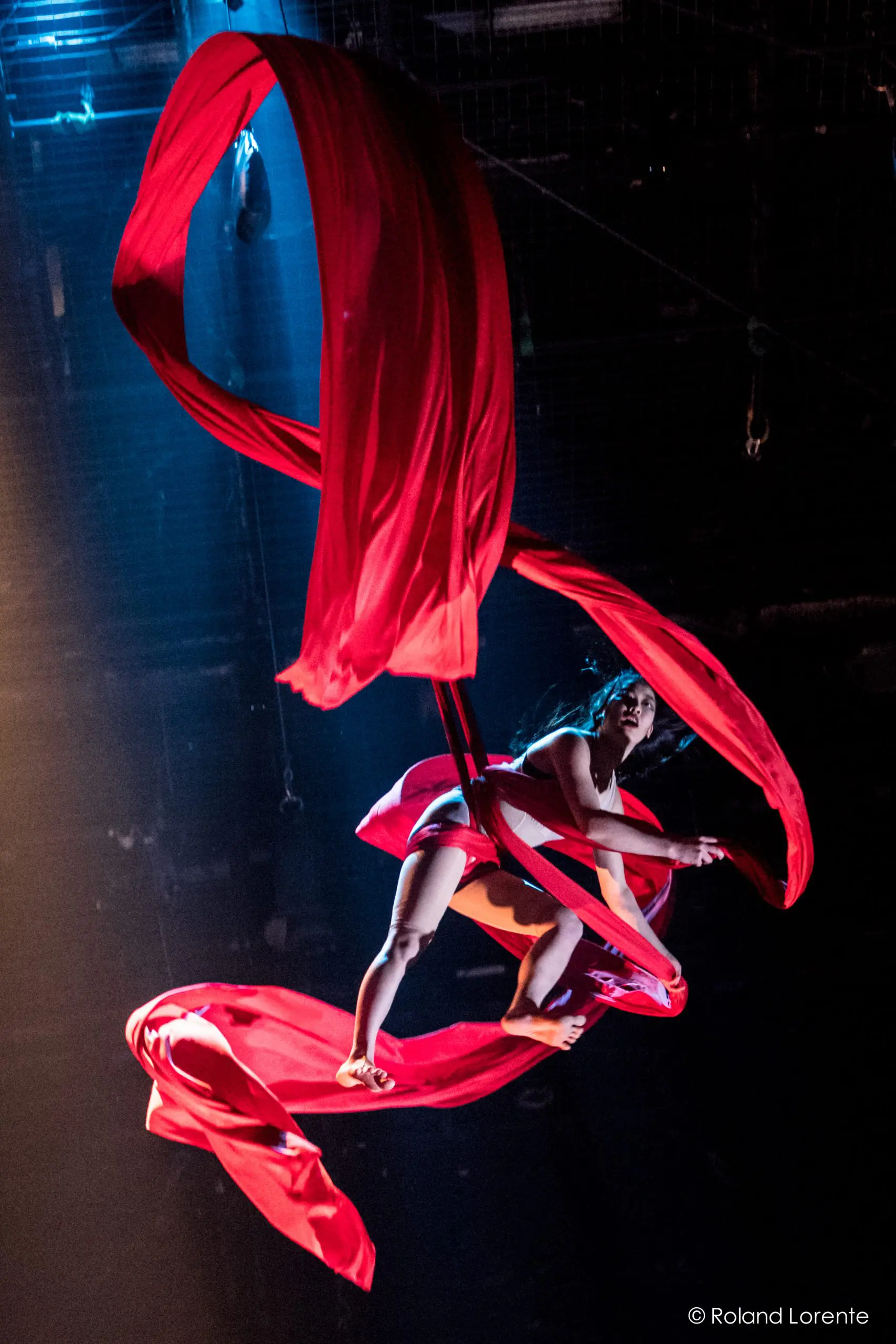 Une artiste aérienne, rappelant Mizuki Shinagawa, est gracieusement suspendue dans les airs, enveloppée dans des rubans de soie rouge fluides, tandis qu'un éclairage de scène dramatique l'illumine sur un fond sombre.