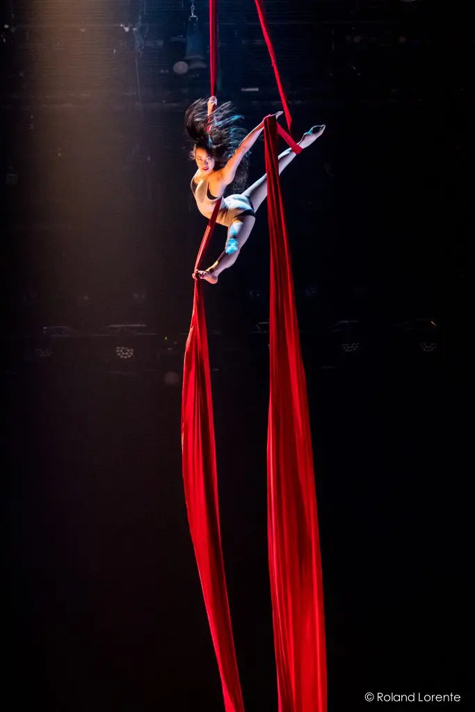 La voltigeuse Mizuki Shinagawa se produit en hauteur, gracieusement suspendue à des rubans de soie rouge, une jambe tendue et les bras en extension, éclairée par les lumières de la scène sur un fond sombre.