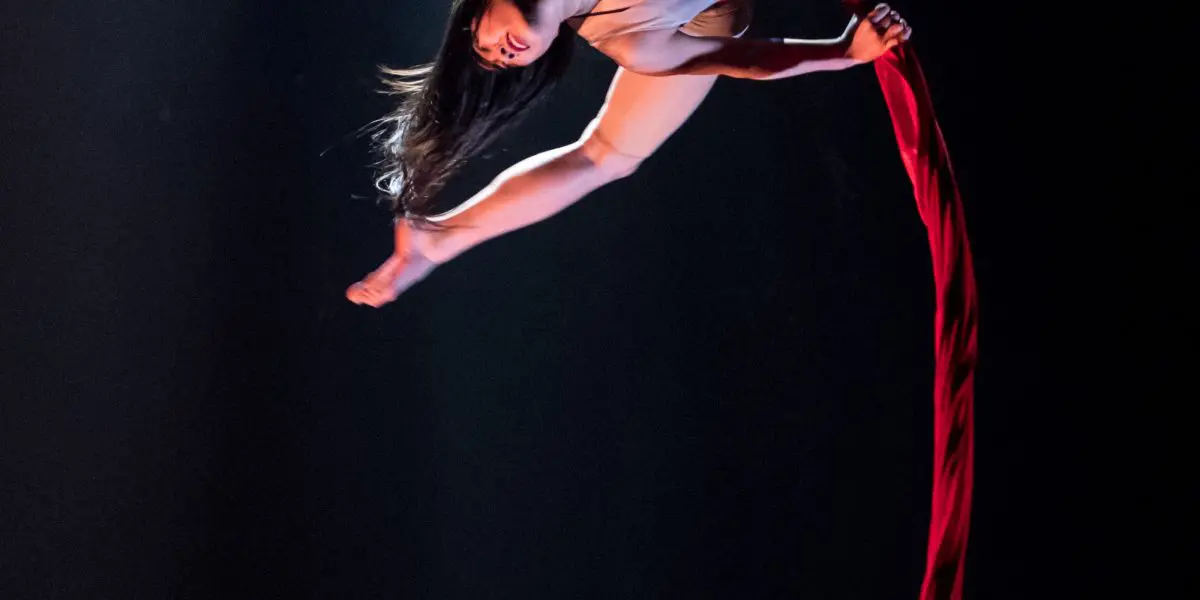 Une femme, Mizuki Shinagawa, exécute un numéro de soie aérienne, suspendue à l'envers par un tissu rouge, une jambe tendue et les cheveux pendants, sur un fond de scène sombre.
