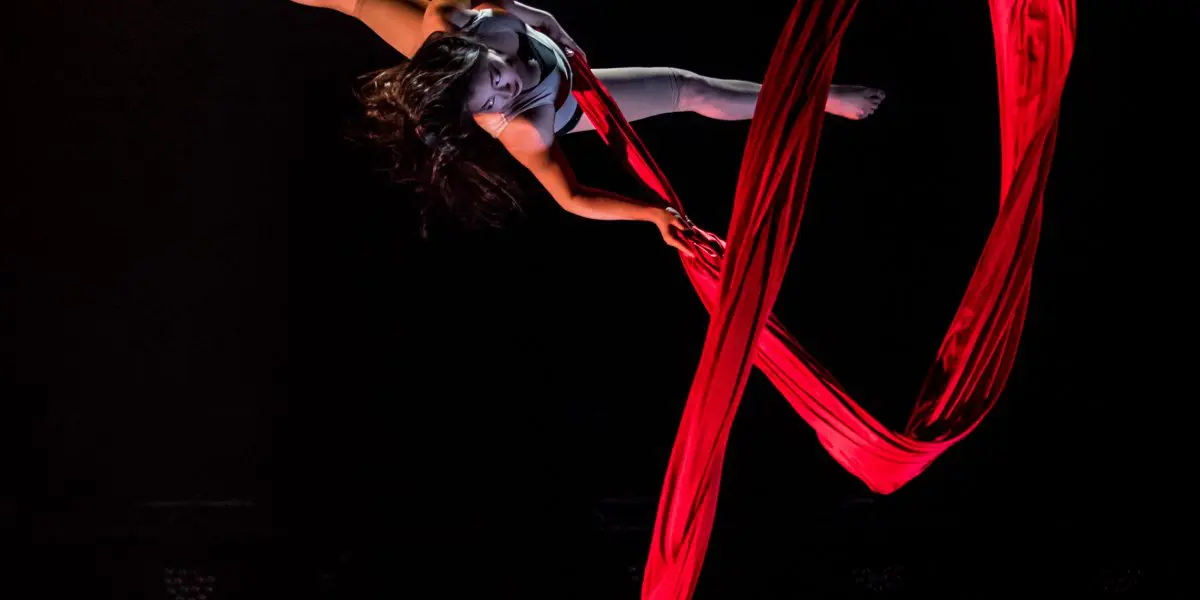 Une voltigeuse, Mizuki Shinagawa, se produit sur des soies rouges, suspendue dans les airs sur un fond sombre, le tissu de soie s'enroulant et s'écoulant élégamment autour d'elle.