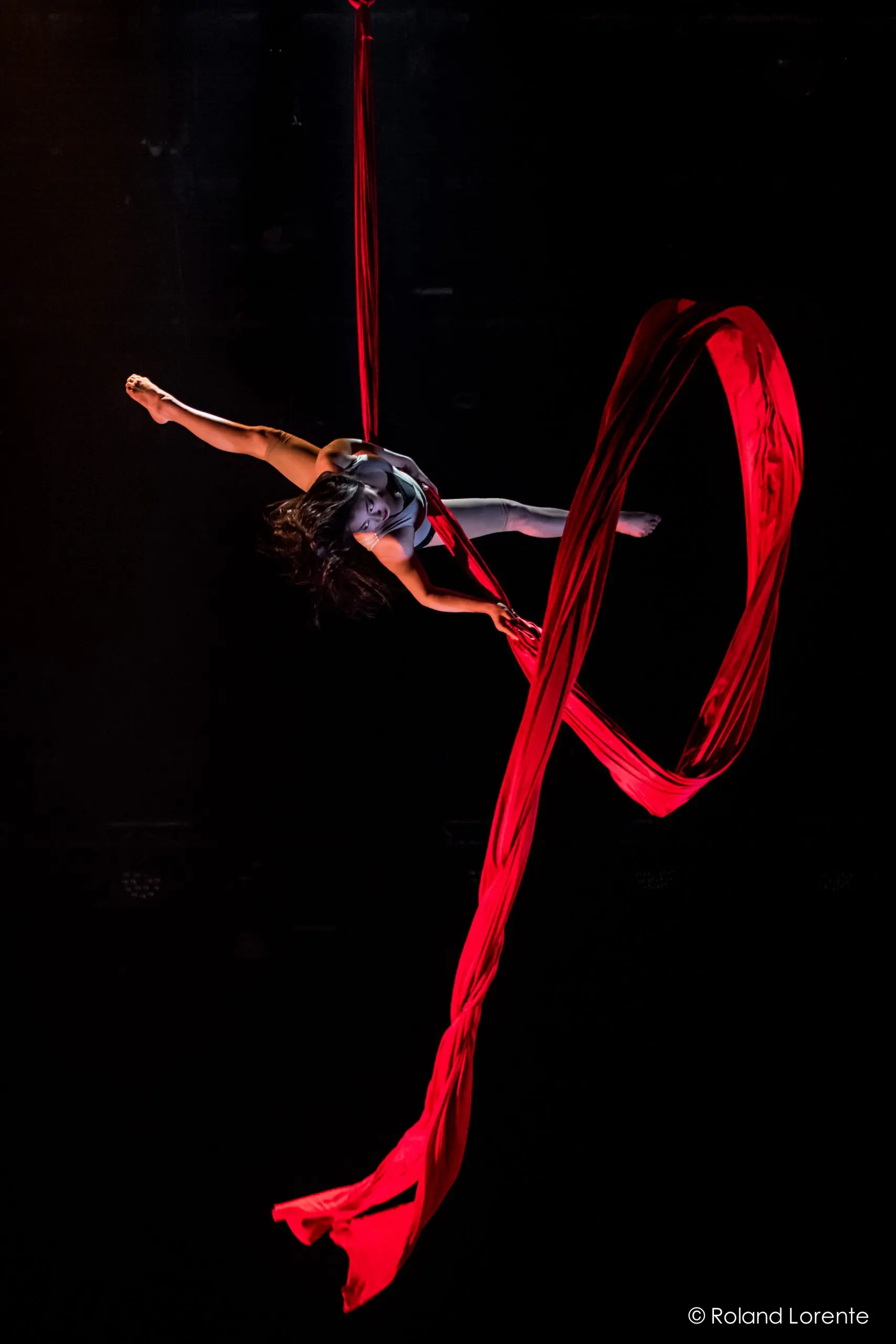 Une voltigeuse, Mizuki Shinagawa, se produit sur des soies rouges, suspendue dans les airs sur un fond sombre, le tissu de soie s'enroulant et s'écoulant élégamment autour d'elle.