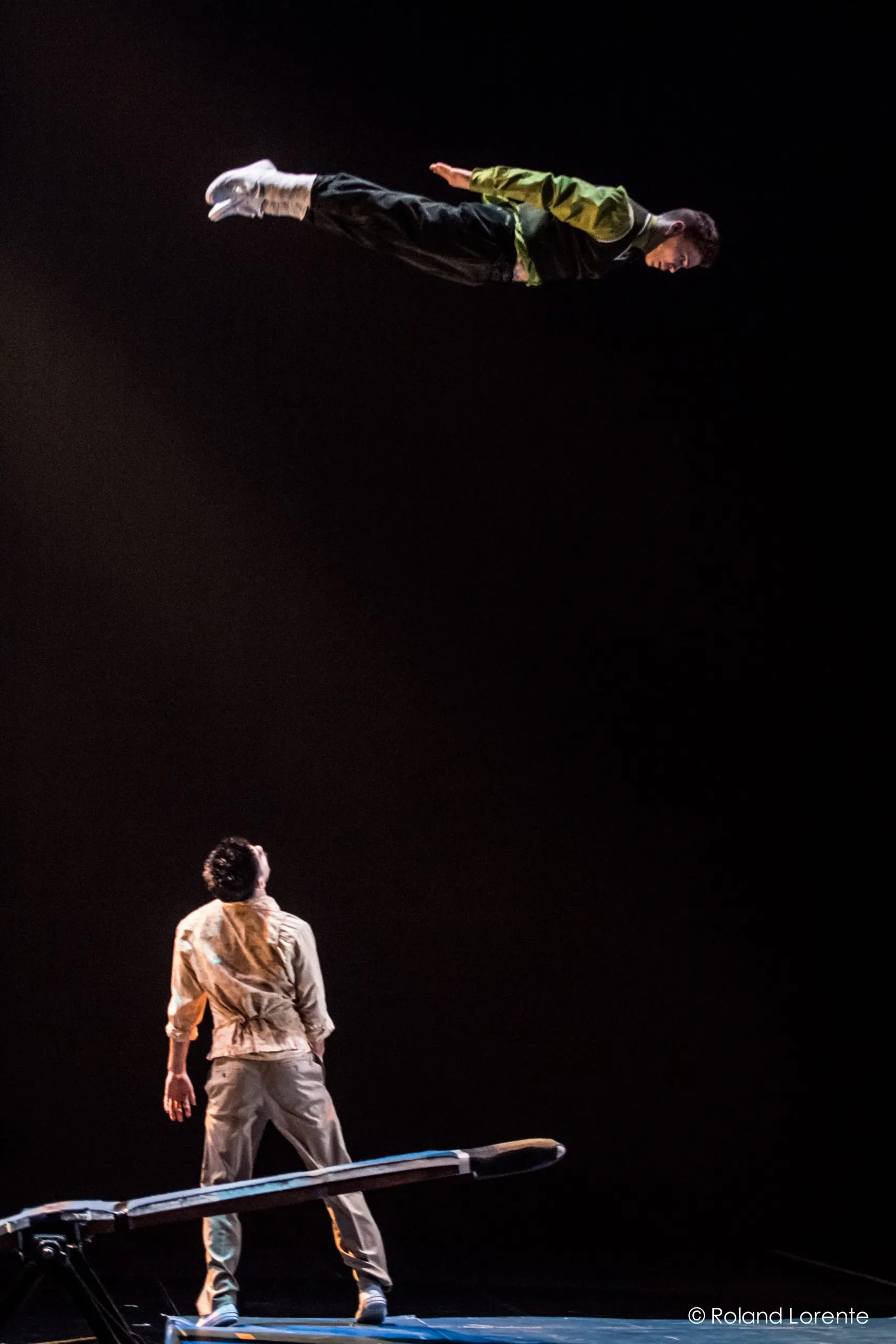 Un interprète en plein vol, lancé d'un appareil à bascule, tandis que Guillaume Larouche se tient en dessous, regardant vers le haut sur une scène sombre.