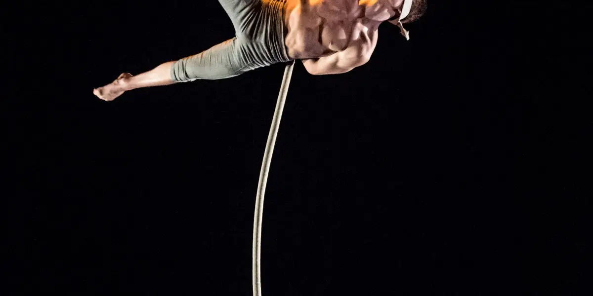 Un acrobate torse nu, Maxime Piché-Luneau, vêtu d'un pantalon gris, exécute un numéro aérien en saisissant une corde verticale d'une main et en tendant la jambe sur un fond sombre. Une corde enroulée est posée sur le sol.