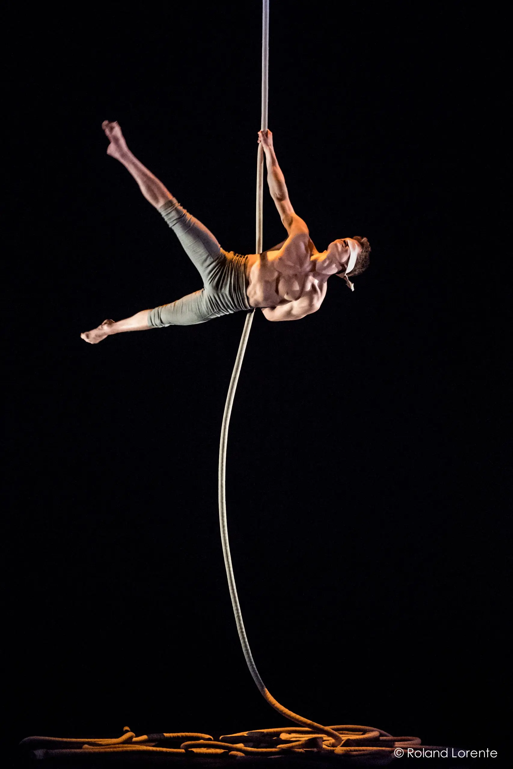 Un acrobate torse nu, Maxime Piché-Luneau, vêtu d'un pantalon gris, exécute un numéro aérien en saisissant une corde verticale d'une main et en tendant la jambe sur un fond sombre. Une corde enroulée est posée sur le sol.