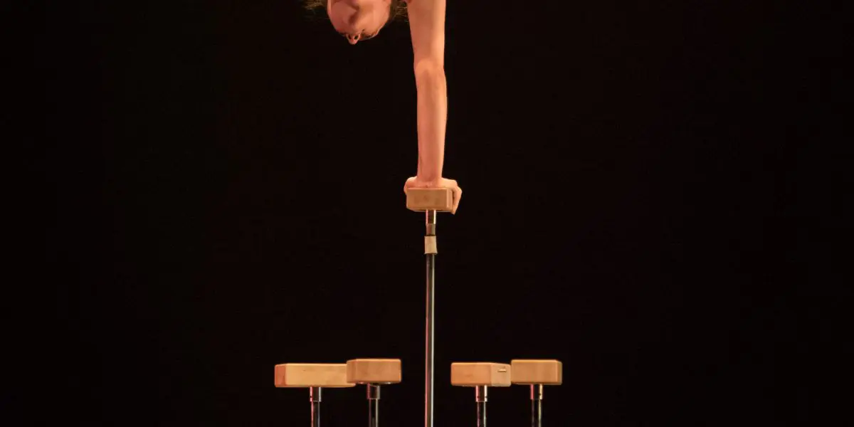 Danielle Saulnier, une acrobate chevronnée, se tient en équilibre sur une main à l'aide de cannes, effectuant un spectaculaire écart avec les jambes étendues au-dessus d'une plate-forme en bois sur un fond sombre.