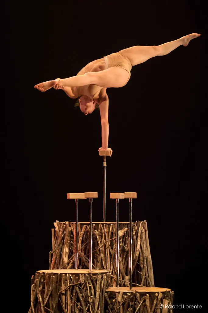 Danielle Saulnier, une acrobate chevronnée, se tient en équilibre sur une main à l'aide de cannes, effectuant un spectaculaire écart avec les jambes étendues au-dessus d'une plate-forme en bois sur un fond sombre.