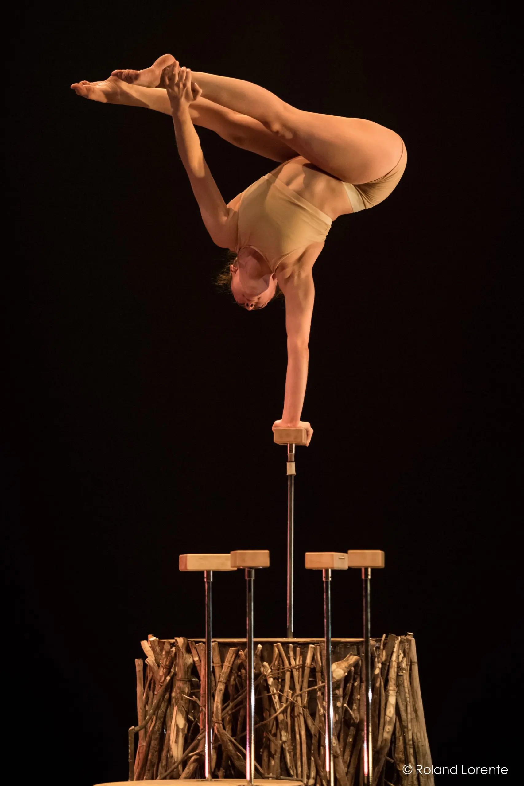 Danielle Saulnier, une acrobate chevronnée, se tient en équilibre sur une main au sommet d'une paire de cannes, les jambes tendues gracieusement au-dessus de la tête. Elle évolue au-dessus d'une base de bâtons en bois disposés sur un fond sombre.