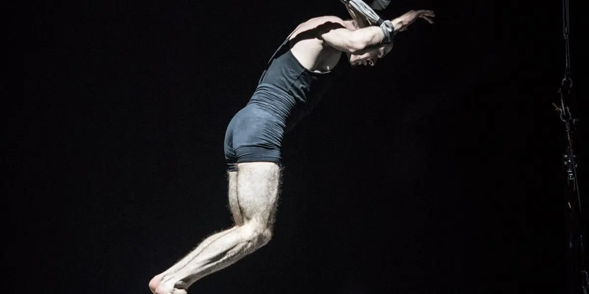 Un artiste, Flavien Marciasini, vêtu d'une tenue noire, se tient en équilibre sur une chaise blanche, penché en avant, les bras levés, tout en étant suspendu par de multiples sangles dans un décor sombre et dramatique.