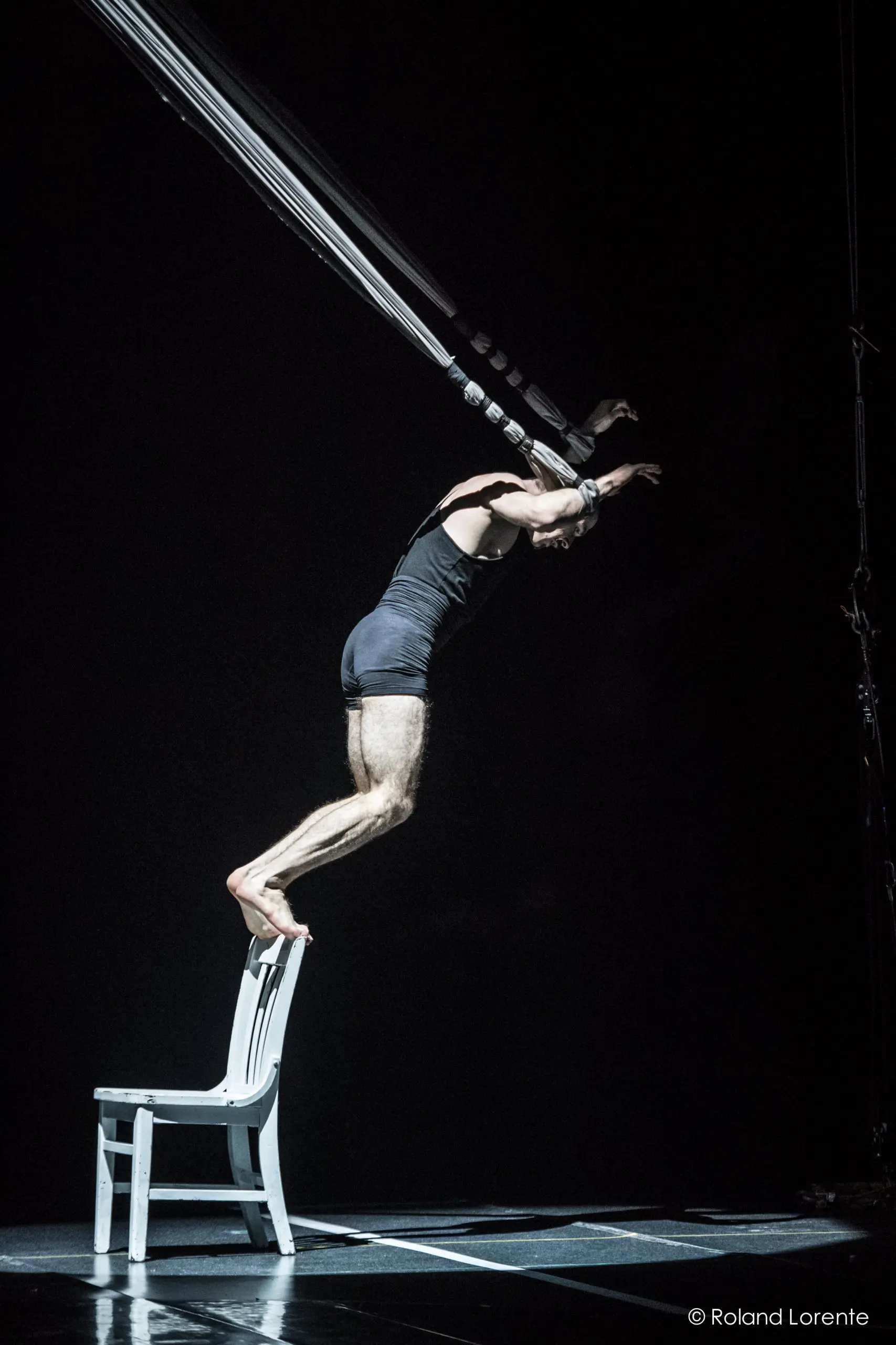 Un artiste, Flavien Marciasini, vêtu d'une tenue noire, se tient en équilibre sur une chaise blanche, penché en avant, les bras levés, tout en étant suspendu par de multiples sangles dans un décor sombre et dramatique.