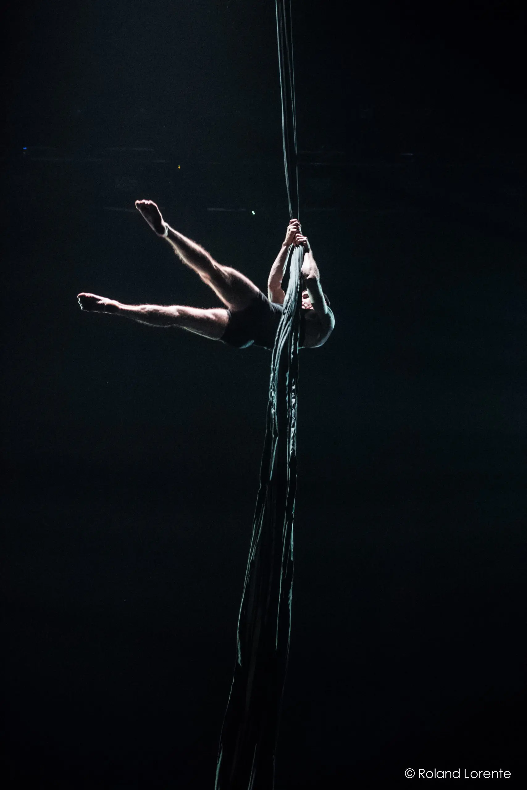 L'artiste aérien Flavien Marciasini se produit sur un long tissu sombre suspendu au plafond, en s'accrochant avec les mains et les jambes tendues, sur un fond sombre et dramatique.