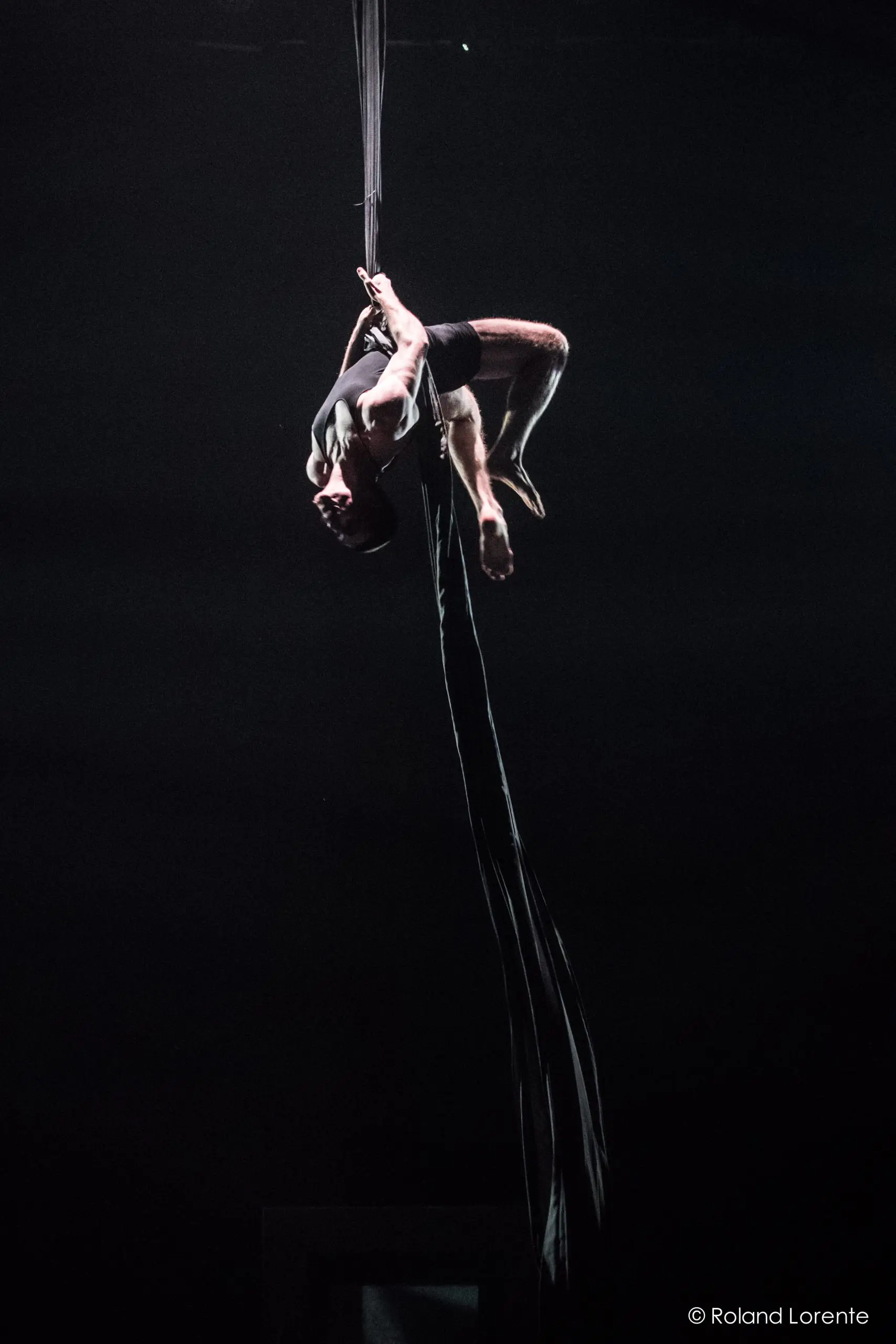 L'artiste aérien Flavien Marciasini exécute une pose à l'envers sur de longues soies noires sur un fond sombre, dramatiquement éclairé par le haut, mettant en valeur une force et un équilibre incroyables.