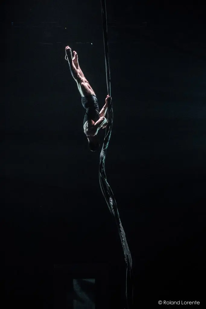 L'artiste aérien Flavien Marciasini exécute un numéro d'acrobatie sur des soies suspendues, la tête en bas, sous un projecteur et sur un fond sombre.