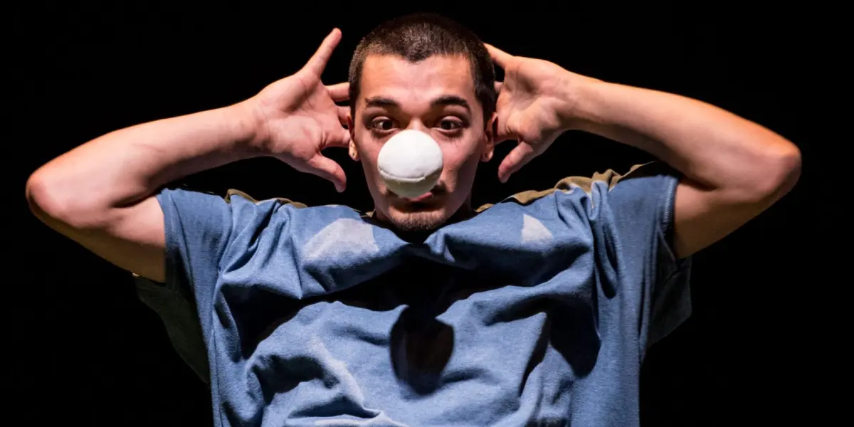 Sereno Aguilar Izzo, vêtu d'une chemise bleue, tient les mains derrière la tête, coudes en l'air, tandis qu'il équilibre une balle blanche sur son nez, sur un fond sombre.