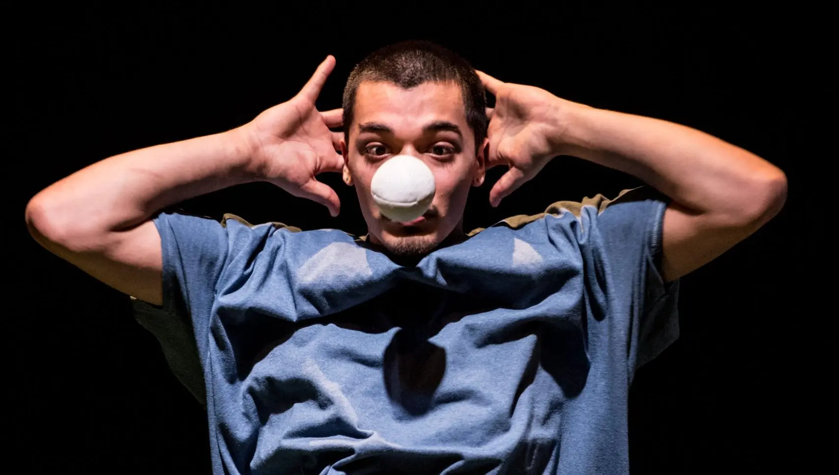 Sereno Aguilar Izzo, vêtu d'une chemise bleue, tient les mains derrière la tête, coudes en l'air, tandis qu'il équilibre une balle blanche sur son nez, sur un fond sombre.