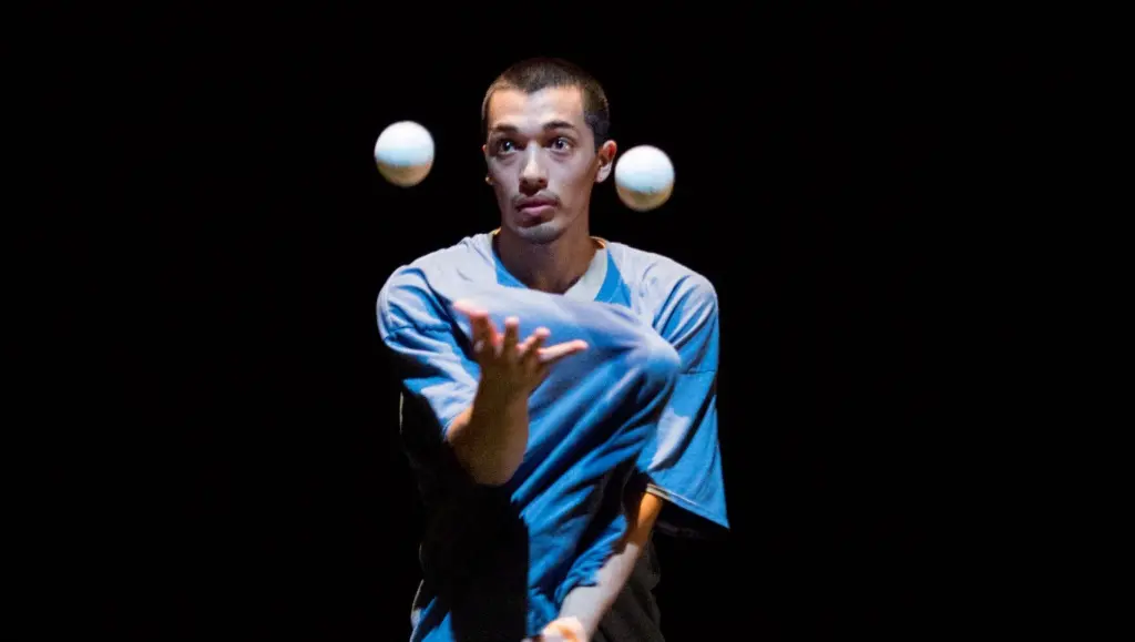 Une personne portant une chemise bleue, rappelant le style de Sereno Aguilar Izzo, jongle avec deux balles blanches sur un fond sombre, en se concentrant sur les objets en mouvement.