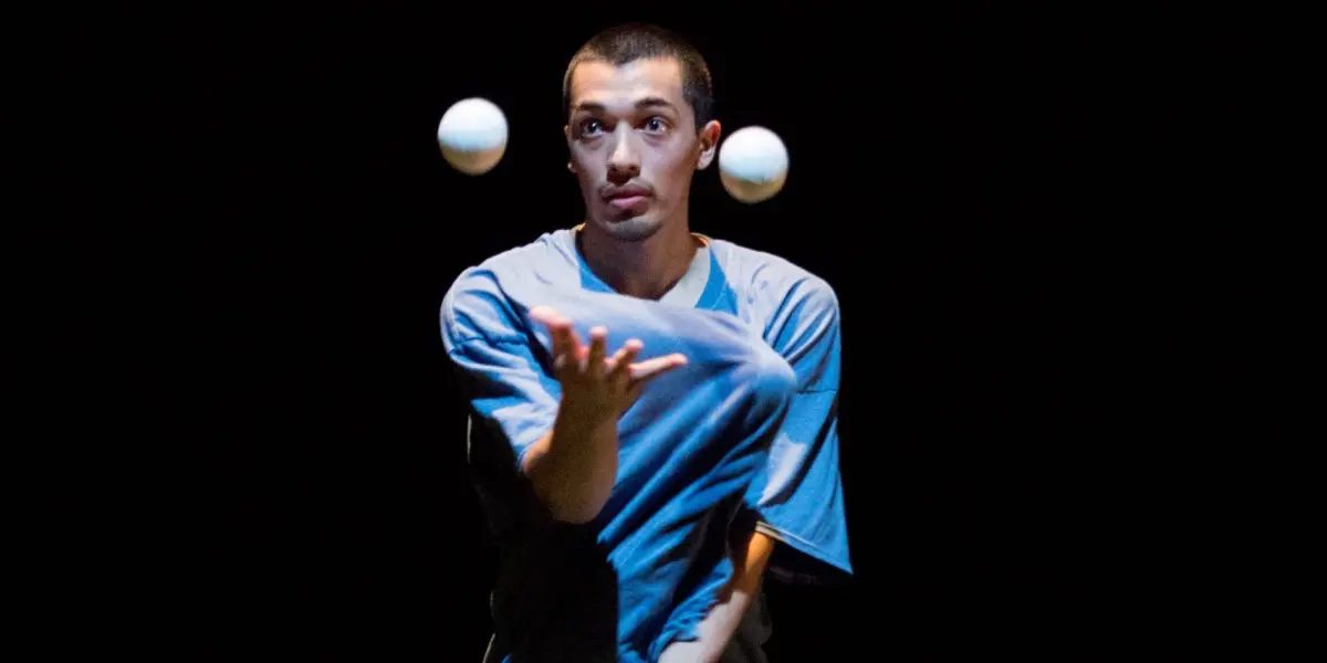Une personne portant une chemise bleue, rappelant le style de Sereno Aguilar Izzo, jongle avec deux balles blanches sur un fond sombre, en se concentrant sur les objets en mouvement.